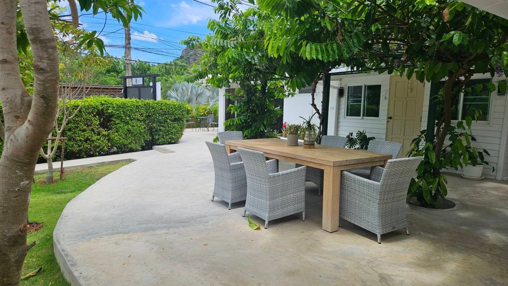 Garden dining table with wicker chairs under shade trees, beside the lawn and the white bungalows at The White Room