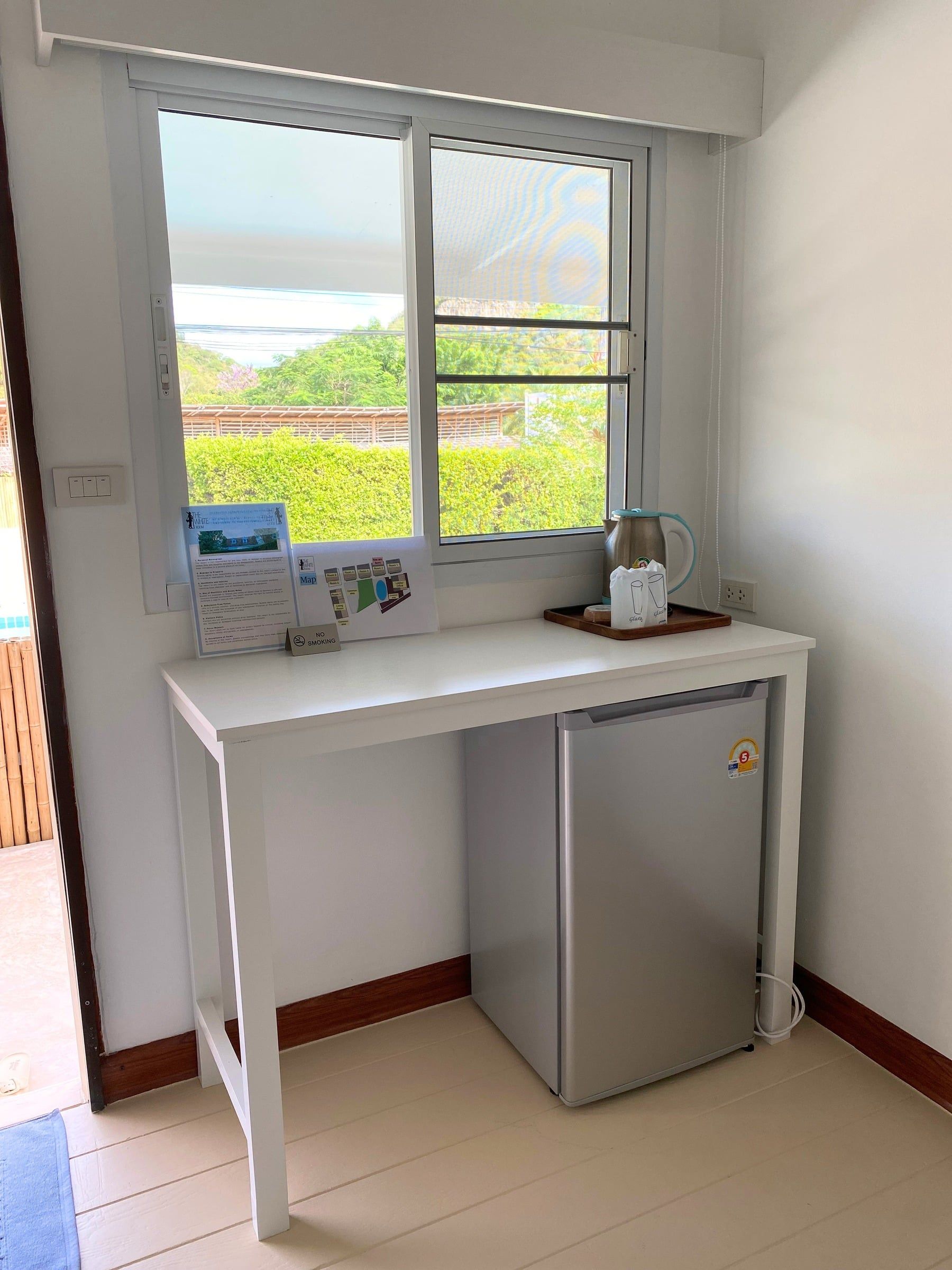 Desk under a window with a mini fridge and kettle, and greenery outside